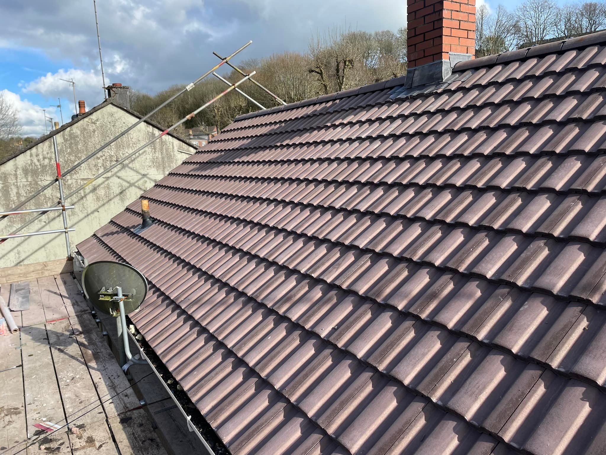 A rooftop with brown tiles under a partly cloudy sky. Scaffolding is visible on the left side, and a satellite dish is mounted near the edge. A red-brick chimney stands near the center. Trees are visible in the background.