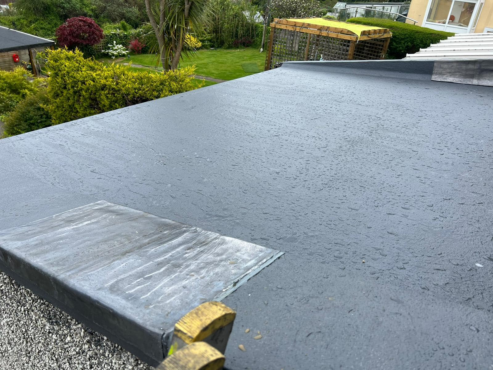 A flat roof with a textured, dark surface showing signs of rain. In the background, there is a lush garden with various shrubs and trees. A light-colored house is partially visible to the right.