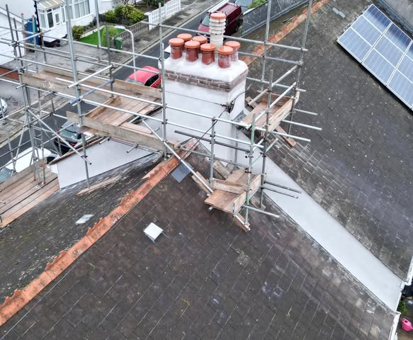 A rooftop under construction with scaffolding surrounding a chimney. Solar panels are visible on one section of the roof. Nearby, buildings and parked cars can be seen from an aerial perspective.