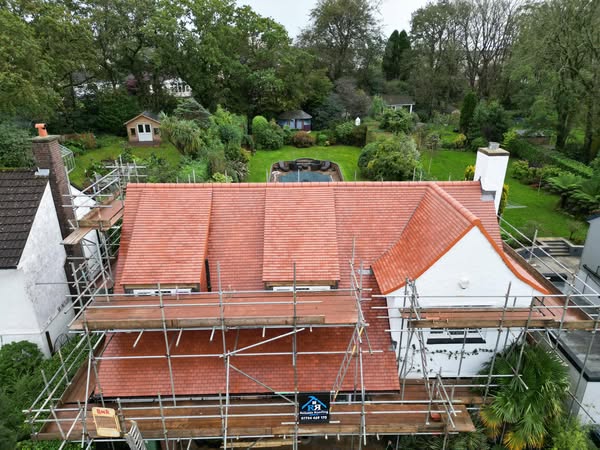 A red-roofed house surrounded by scaffolding, set in a lush green garden with trees and shrubs. A small shed and a pool are visible in the backyard, under an overcast sky.