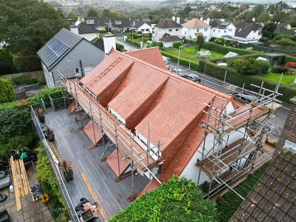 Aerial view of a house under renovation with red-tiled roof and scaffolding on both sides. The house is surrounded by green hedges, nearby houses, and a road. Solar panels are visible on a nearby building. Weather is overcast.