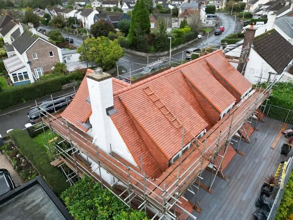 Aerial view of a steep-roofed white house under construction. The roof is newly tiled with red tiles and surrounded by scaffolding. Nearby are houses, trees, and a road with parked cars on a cloudy day.