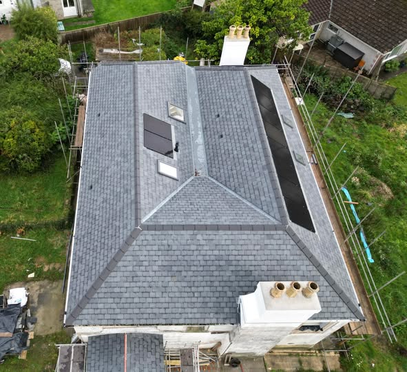 Aerial view of a house with a grey slate roof, featuring skylights and chimneys. Scaffolding surrounds the building, and there are green lawns and trees visible around the property.