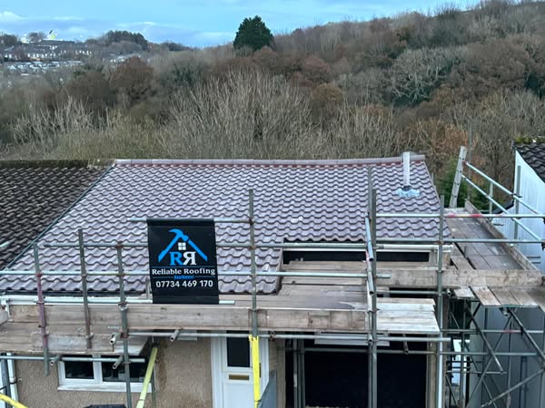 A house with a new, tiled roof and scaffolding in front. A sign on the scaffolding reads "Reliable Roofing" with a phone number. Trees and a hilly landscape are visible in the background.