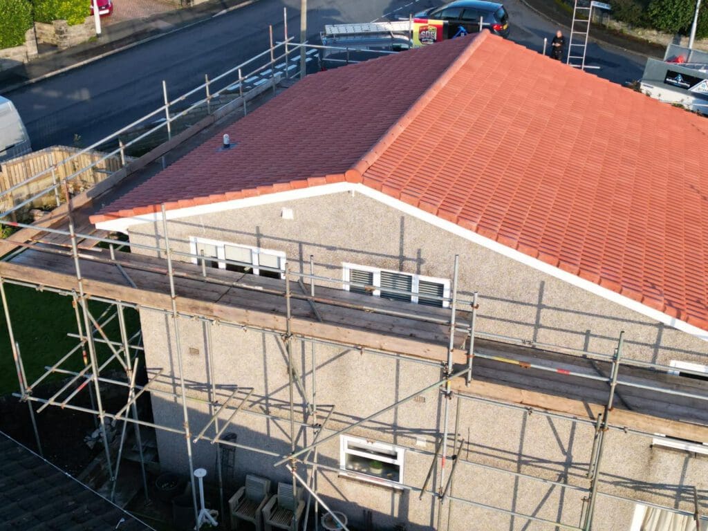 Aerial view of a house with a red-tiled roof and surrounding scaffolding. The building appears to be under construction or renovation. There are roads and parked vehicles nearby. The setting is residential.
