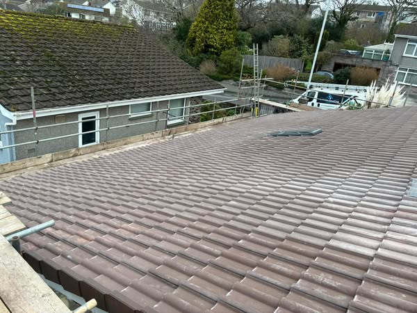 View of a residential rooftop with brown tiles, surrounded by scaffolding. Nearby, a small skylight is visible on the roof. In the background, there are houses, trees, and vehicles parked on the street.