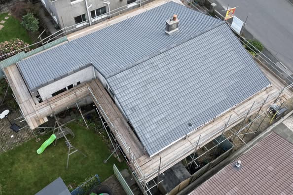 Aerial view of a house with a grey tiled roof under construction. Scaffolding surrounds the house, and a small garden with a slide and swing set is visible. Nearby is a road and neighboring buildings.