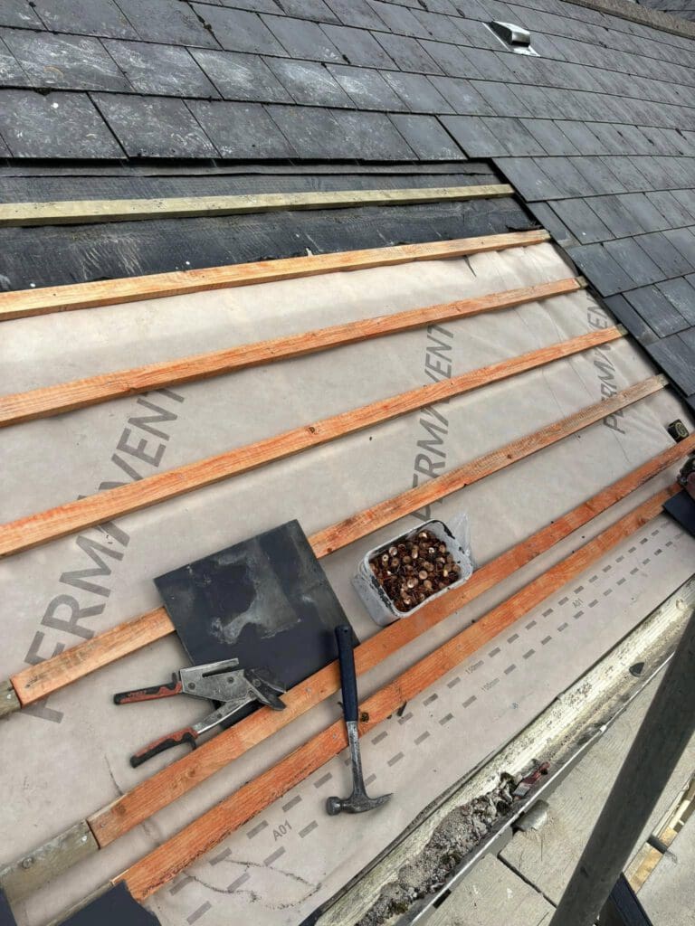 Roof under construction with wooden battens on felt paper. A hammer, two pliers, and roofing nails in a tray are visible. Partially installed slate tiles are on the left.