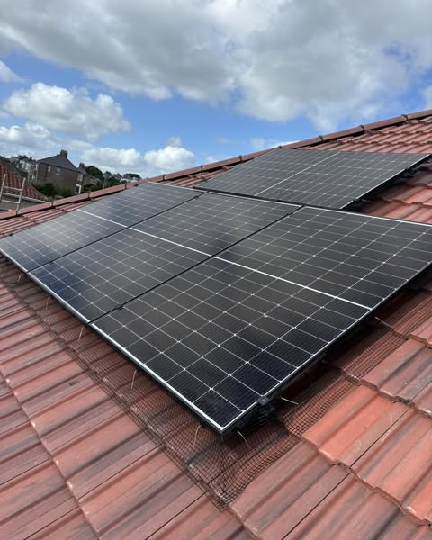 Solar panels installed on a sloped red-tiled roof under a partly cloudy sky. In the background, there are residential buildings and trees.