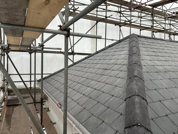 A rooftop covered in gray slate tiles is supported by scaffolding all around. The sky is overcast, and protective netting is visible, suggesting construction or renovation work.