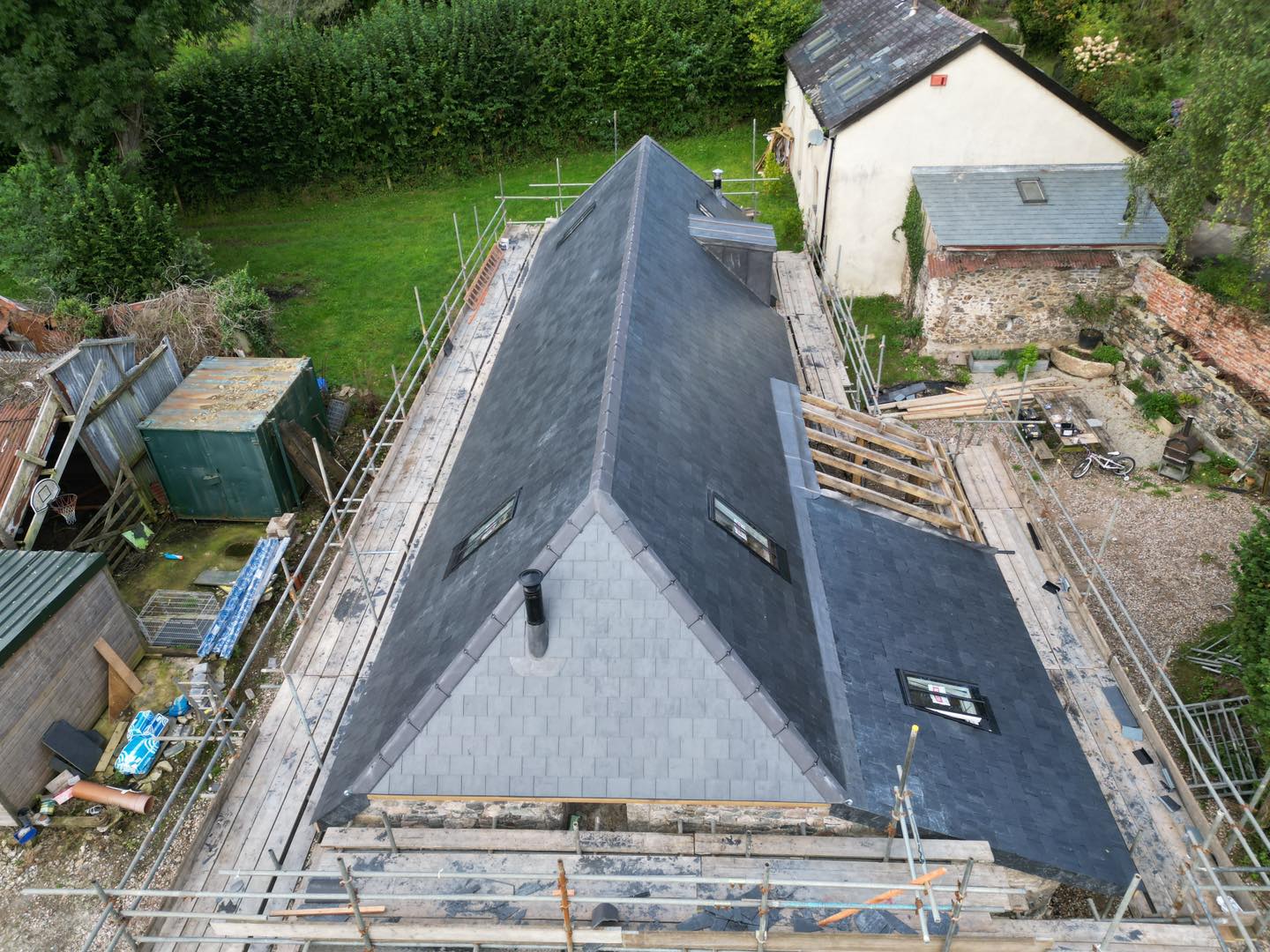 Aerial view of a slate-roofed building under renovation, surrounded by scaffolding. The building is set in a grassy area with trees and another smaller building nearby. Various construction materials are visible on the ground.