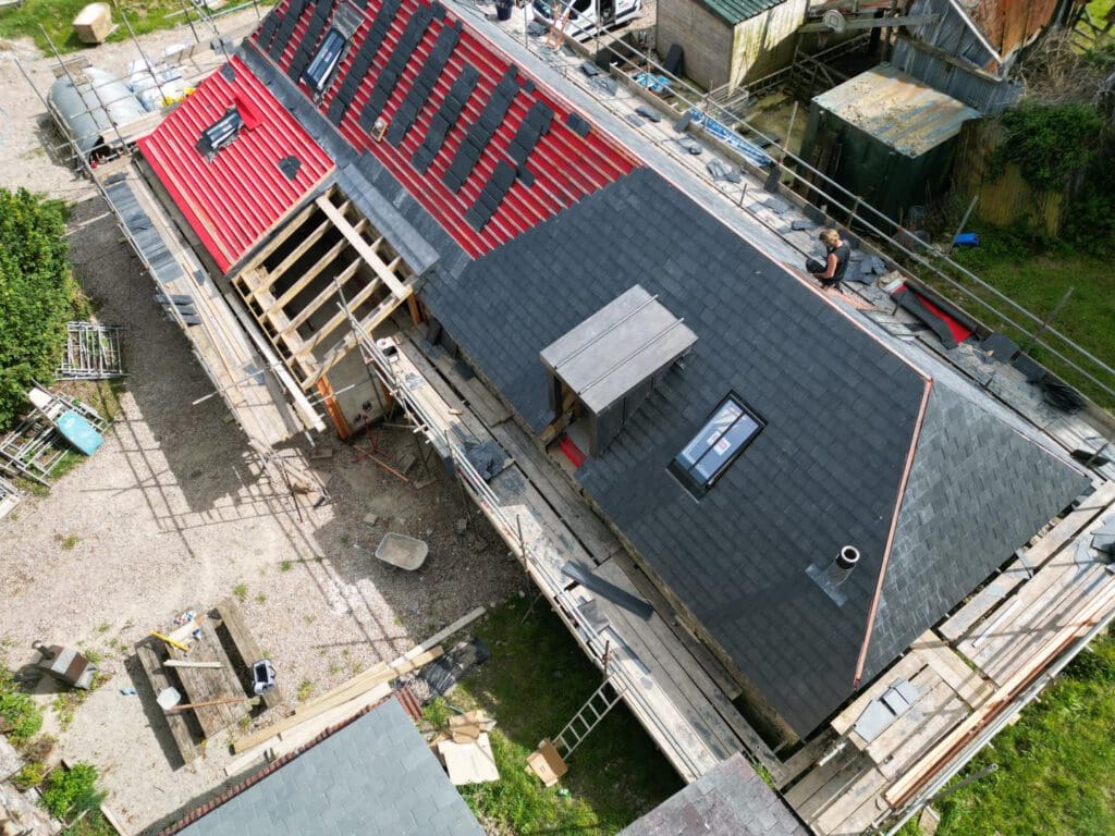Aerial view of a construction site with a long building featuring black and red roofing. Scaffolding surrounds the building, and several ladders are visible. Various construction materials and tools are scattered around the area.