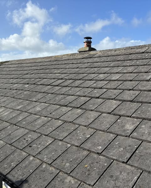 A sloped roof with gray slate tiles under a blue sky with fluffy white clouds. A small chimney with a cap is visible near the ridge of the roof.