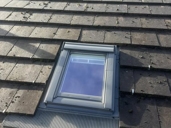A small rectangular skylight is installed on a dark gray slate roof, surrounded by roofing materials. The clear blue sky is reflected in the skylight's glass.