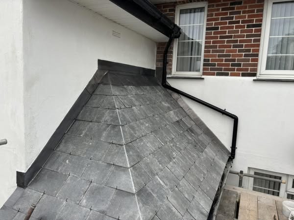 A sloped slate roof with metal flashing connects to a white wall. A black downspout runs from the roof edge to the wall. The building exterior includes a section of red brick with two windows.