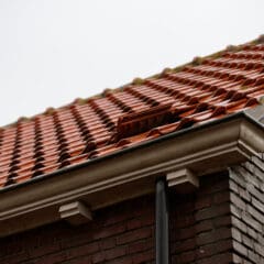Close-up of a rooftop with red clay tiles, some of which are slightly displaced. The roof edges have white wooden detailing, and a rain gutter is seen alongside a brick wall under an overcast sky.