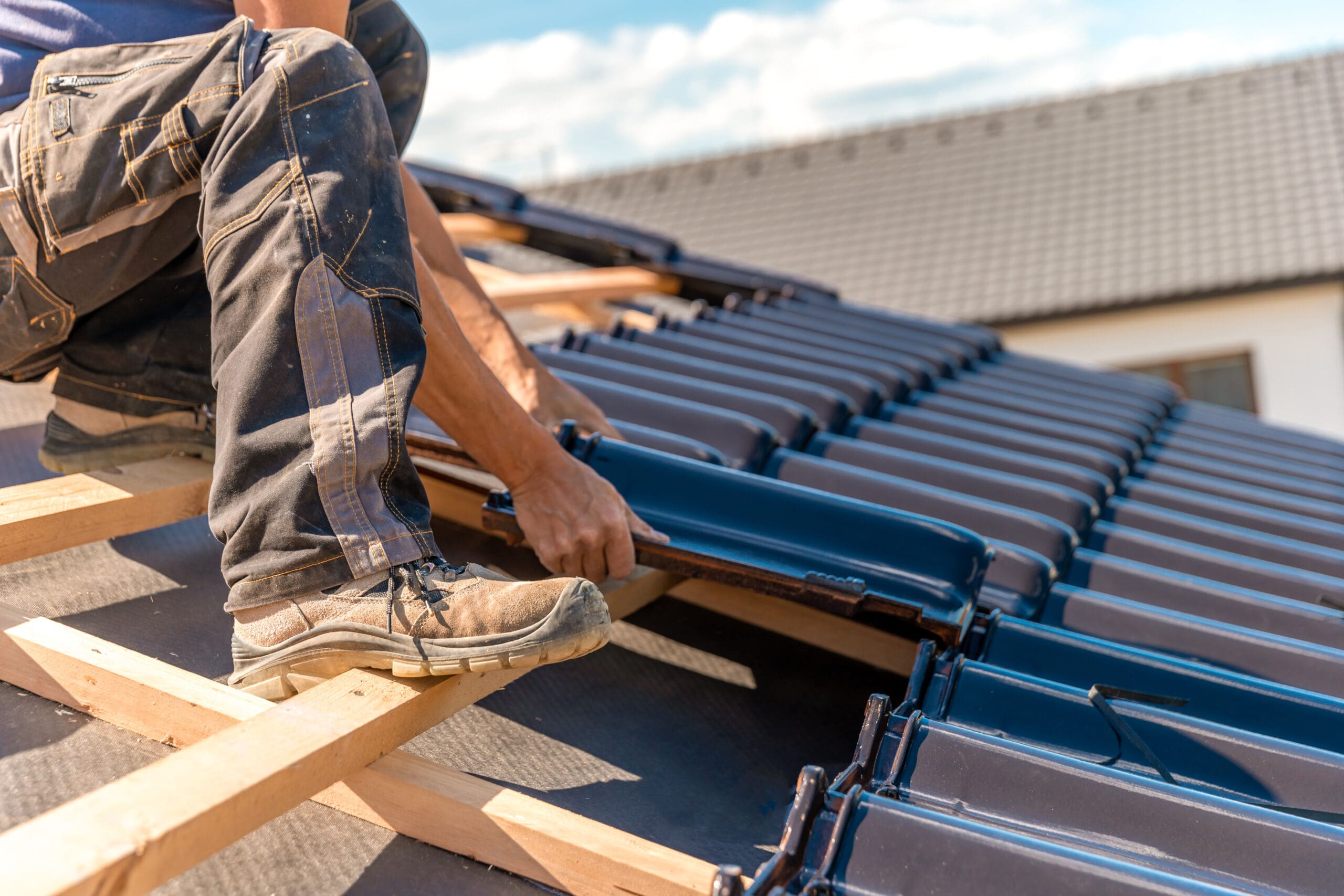 A person wearing work boots installs blue ceramic tiles on a sloped roof under a clear sky. The worker is kneeling and aligning tiles on the wooden framework, with a partially completed roof section visible in the background.