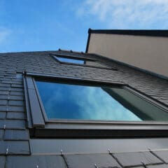 The image shows an upward view of a modern building with a sloped roof covered in dark tiles. Large reflective windows are visible, mirroring a bright blue sky with scattered clouds. The structure has a geometric and sleek design.