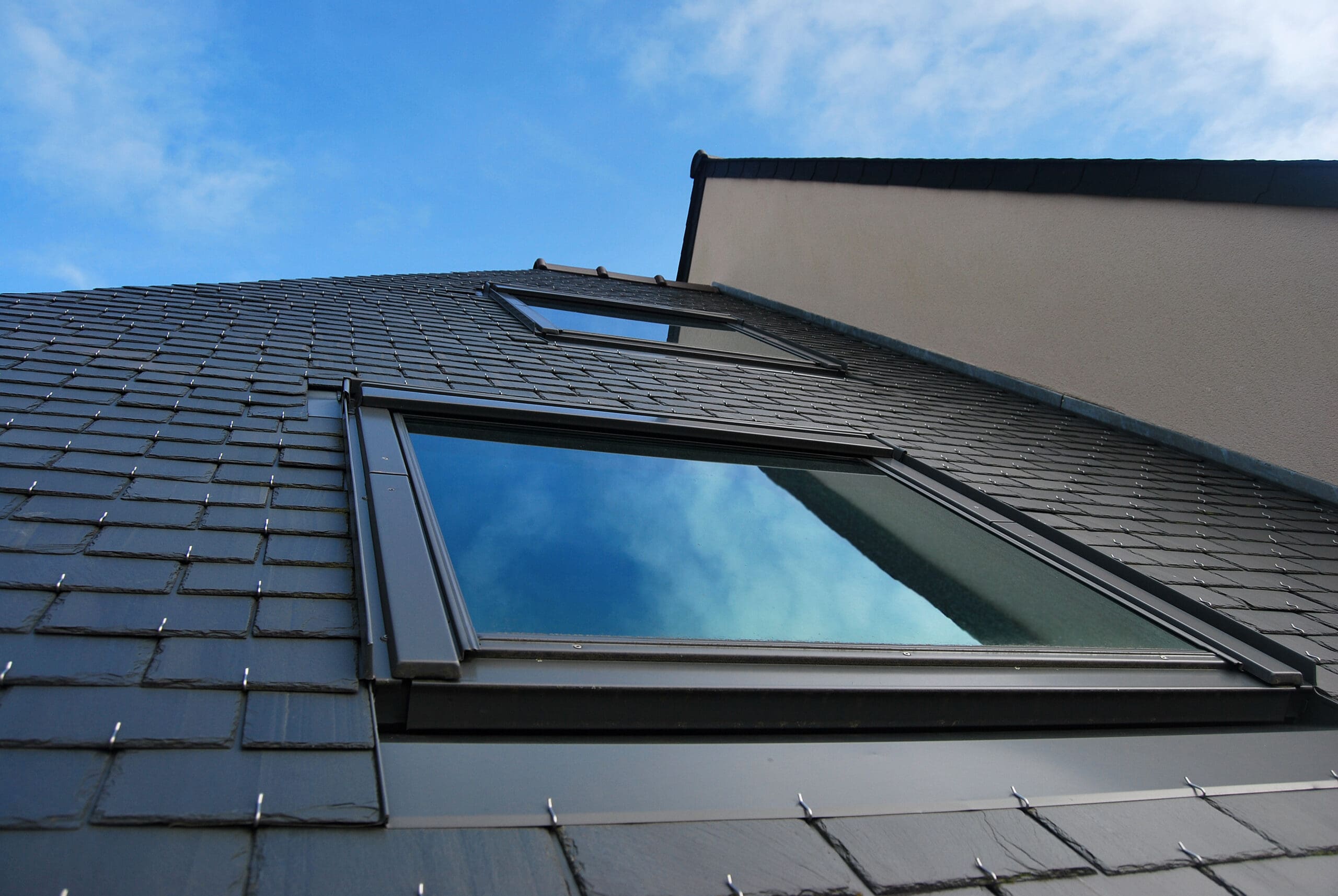The image shows an upward view of a modern building with a sloped roof covered in dark tiles. Large reflective windows are visible, mirroring a bright blue sky with scattered clouds. The structure has a geometric and sleek design.