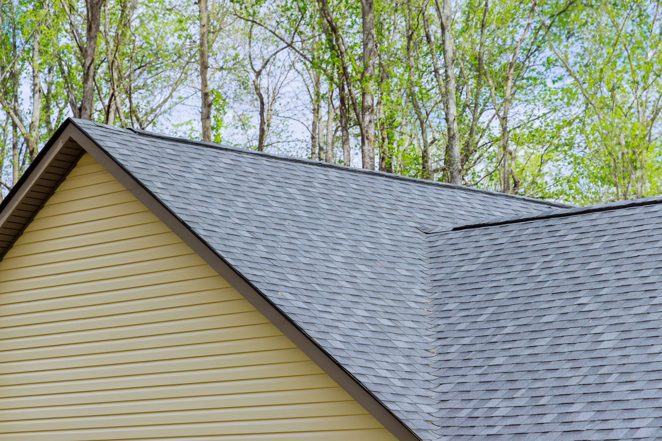 A close-up view of the roof of a house with gray asphalt shingles. The house has yellow siding and is surrounded by tall trees with green foliage under a clear blue sky.
