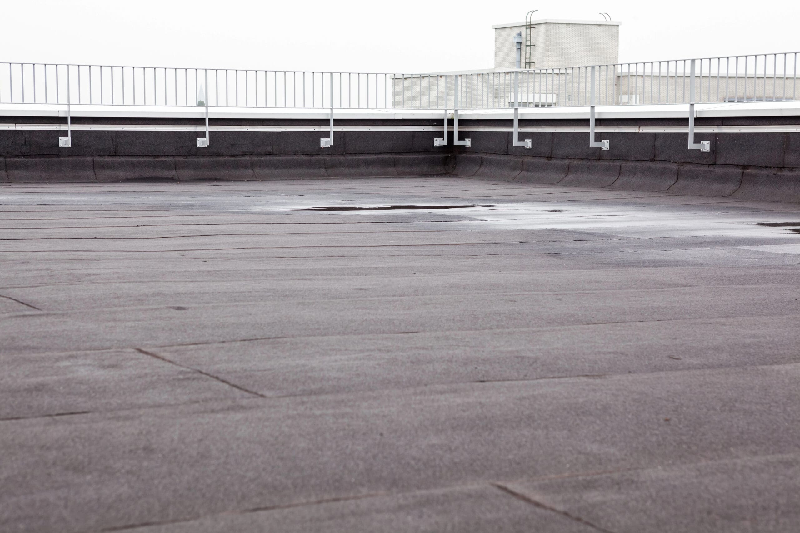 Flat rooftop with a smooth, dark surface, surrounded by a safety railing. A small, light-colored building is visible in the background under an overcast sky. The rooftop has minor puddles, suggesting recent rain.