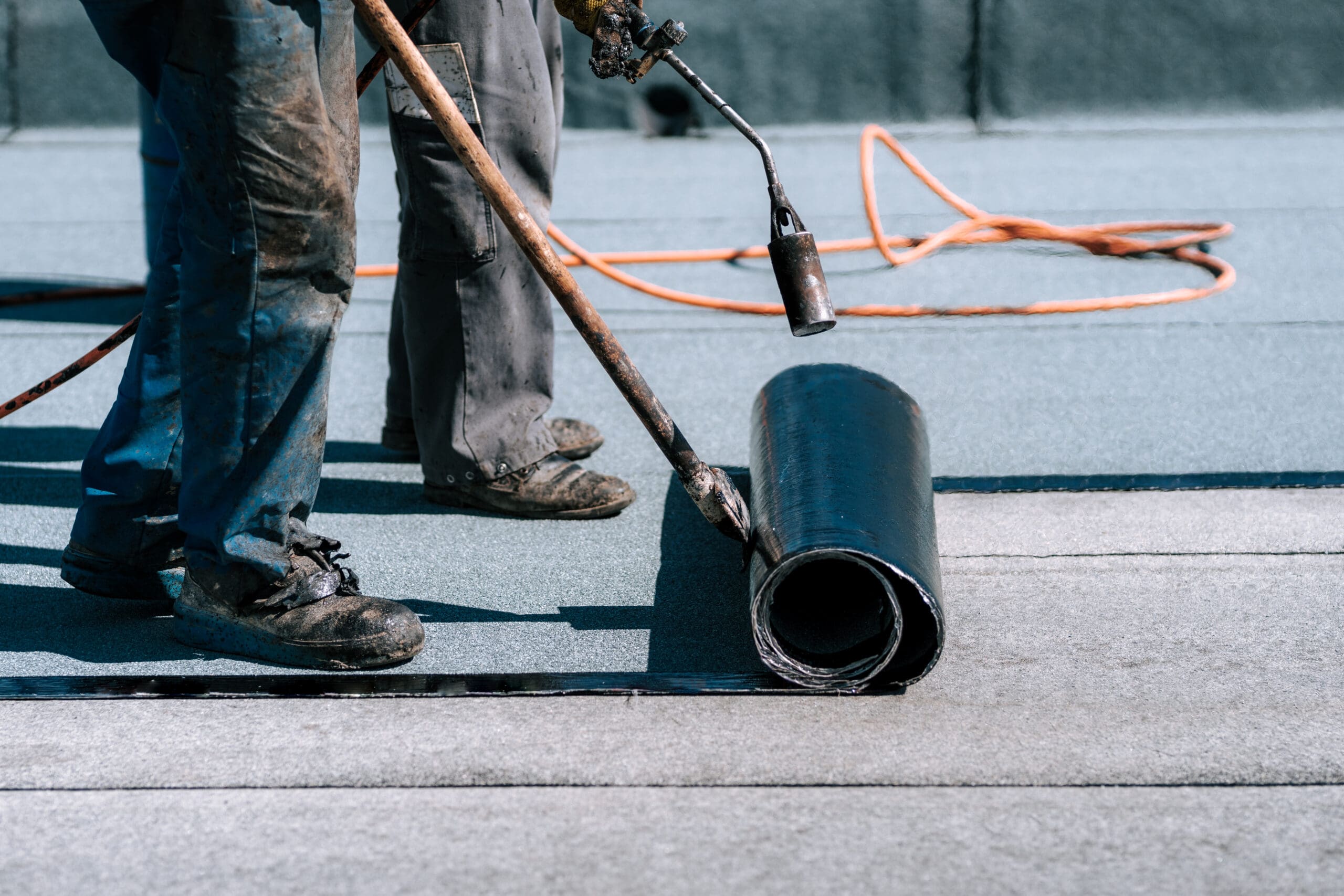 A worker in worn boots and pants uses a torch to seal a roll of black roofing material onto a flat surface. An orange extension cord is visible in the background.