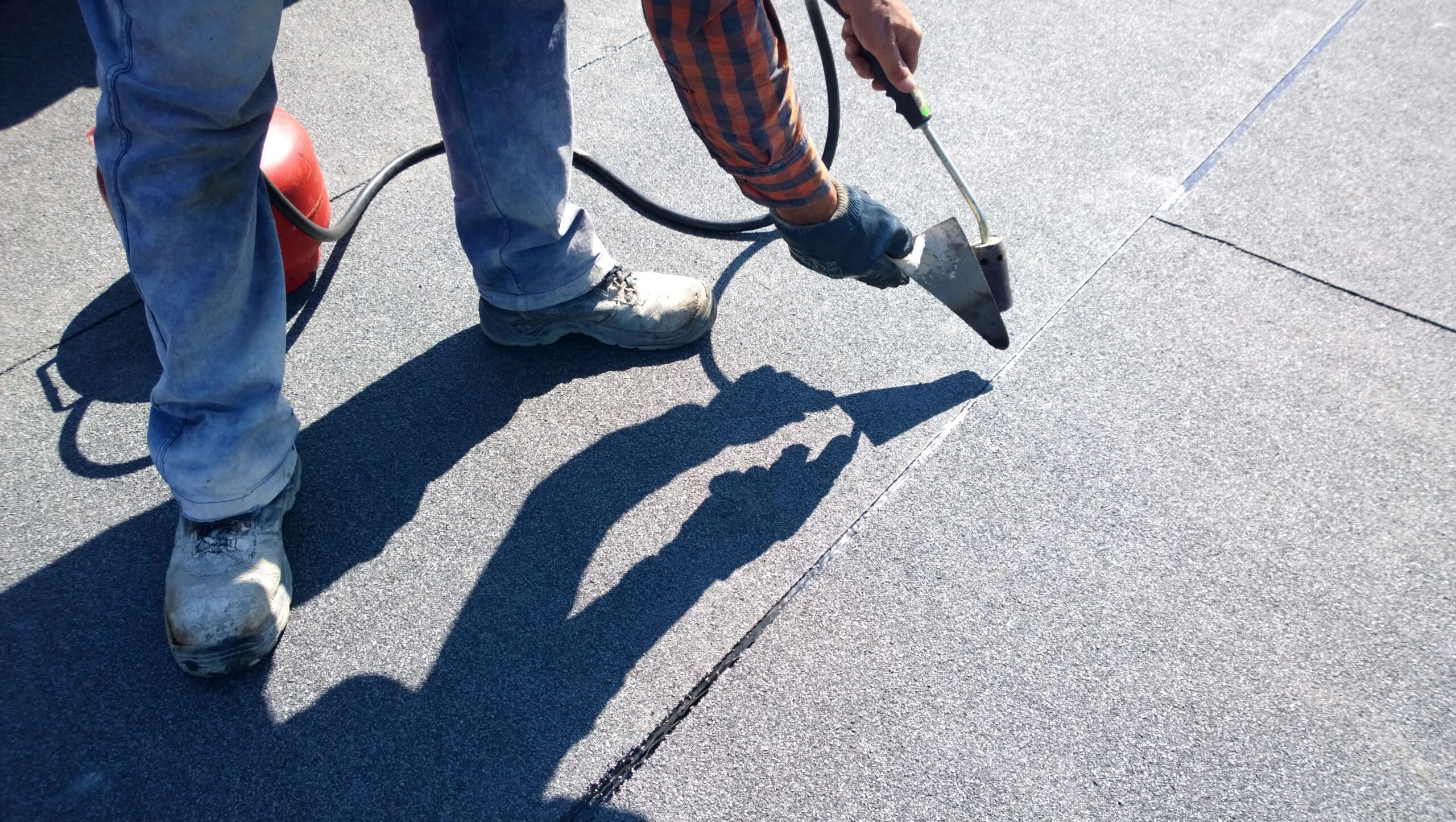A person wearing jeans, boots, and a plaid shirt is using a heat welding tool on a flat, gray roof surface. A red gas tank is nearby on the left. The person's shadow is clearly visible on the roof.
