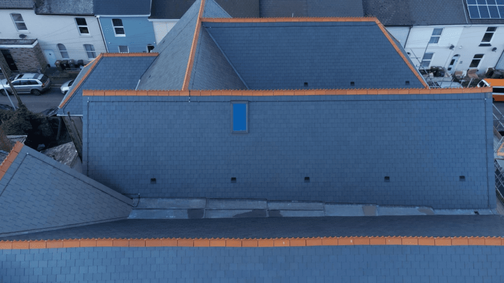 Aerial view of a row of rooftops with dark blue-gray shingles and orange ridge tiles. A single small dormer window is visible on one roof. Several houses and a street can be seen in the background.