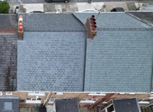 Aerial view of two adjacent tiled roofs, one with red chimneys and the other with gray tiles and four black chimneys. Below, a street with parked cars and a pedestrian sidewalk is visible.