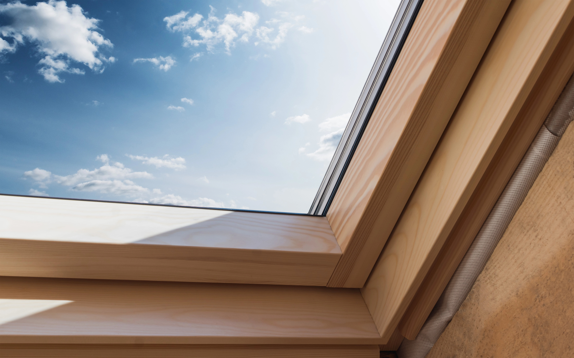 A view of a clear blue sky with a few clouds, seen through a skylight with a wooden frame. The sunlight casts a shadow on the frame, highlighting its texture.