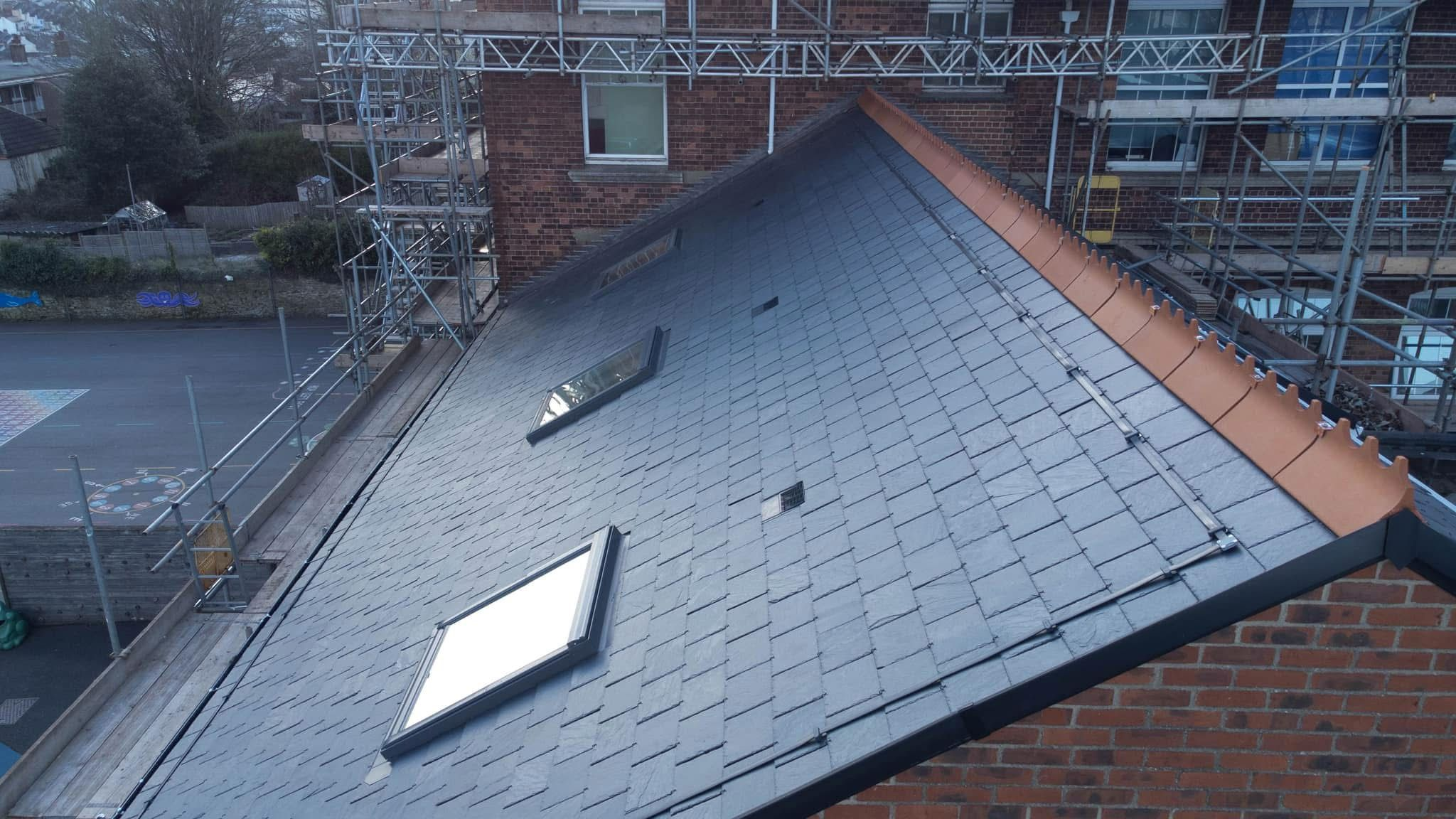 A newly constructed sloped roof with dark shingles and two skylights. The roof is bordered by scaffolding, and part of a brick building is visible. The scene is set on a cloudy day, with a street and additional buildings in the background.